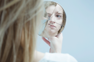 a young woman looks at her reflection in a broken mirror while thinking about bipolar disorder treatment